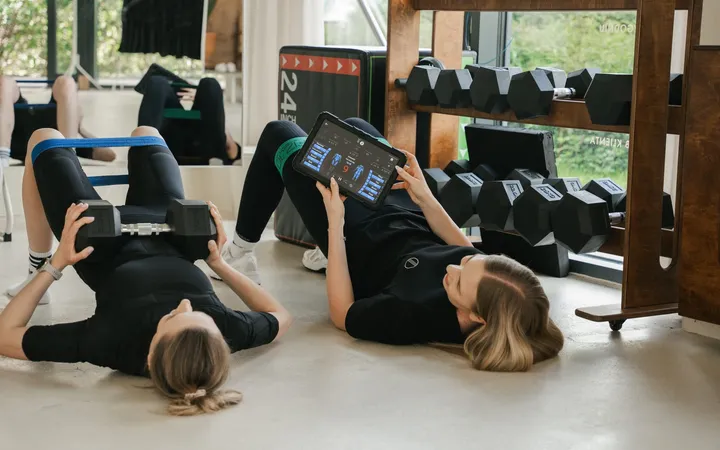 Dwie osoby ćwiczą na macie w studiu WorkItEMS, trenerka pokazuje ćwiczenie na tablecie