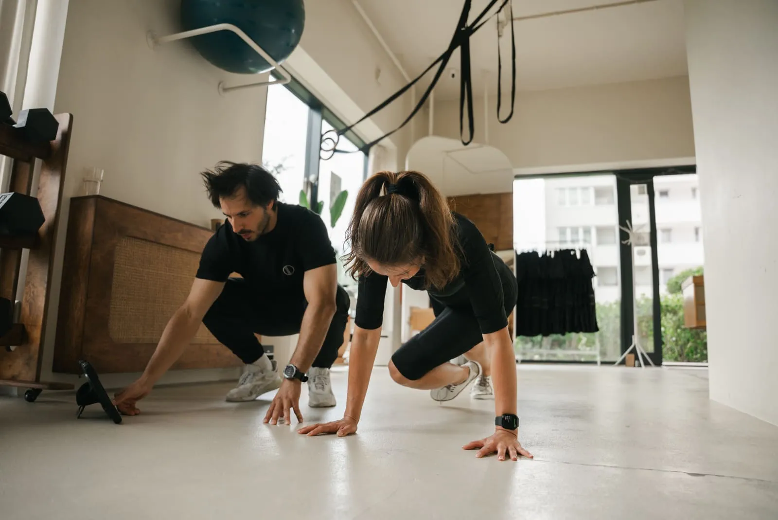 Trener asekuruje klientkę w stroju EMS podczas ćwiczenia w podporze przy oknie