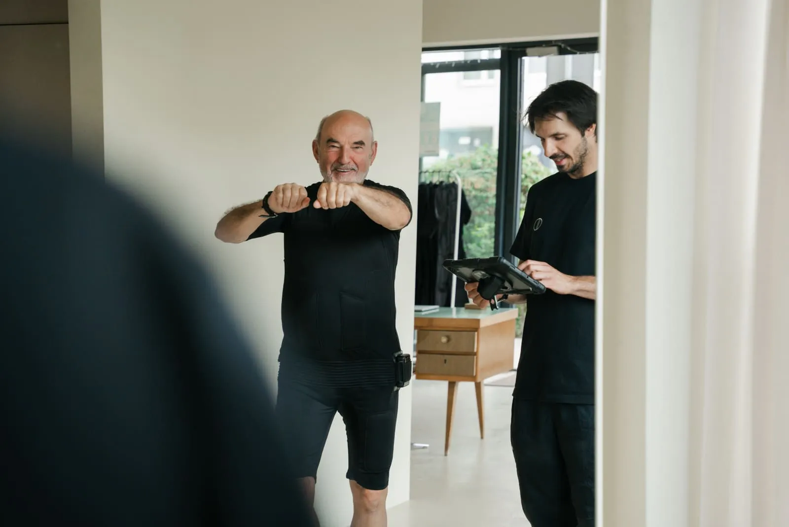 Starszy klient w stroju EMS wykonuje ćwiczenie, trener kontroluje sesję