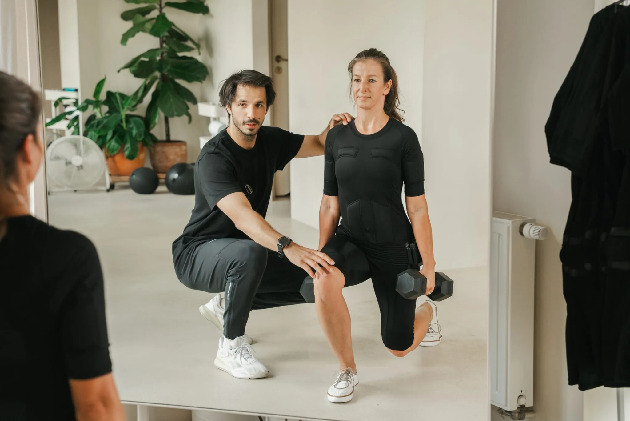Trener asekuruje klientkę w stroju EMS podczas wykroku z hantlami, kadr od lustra