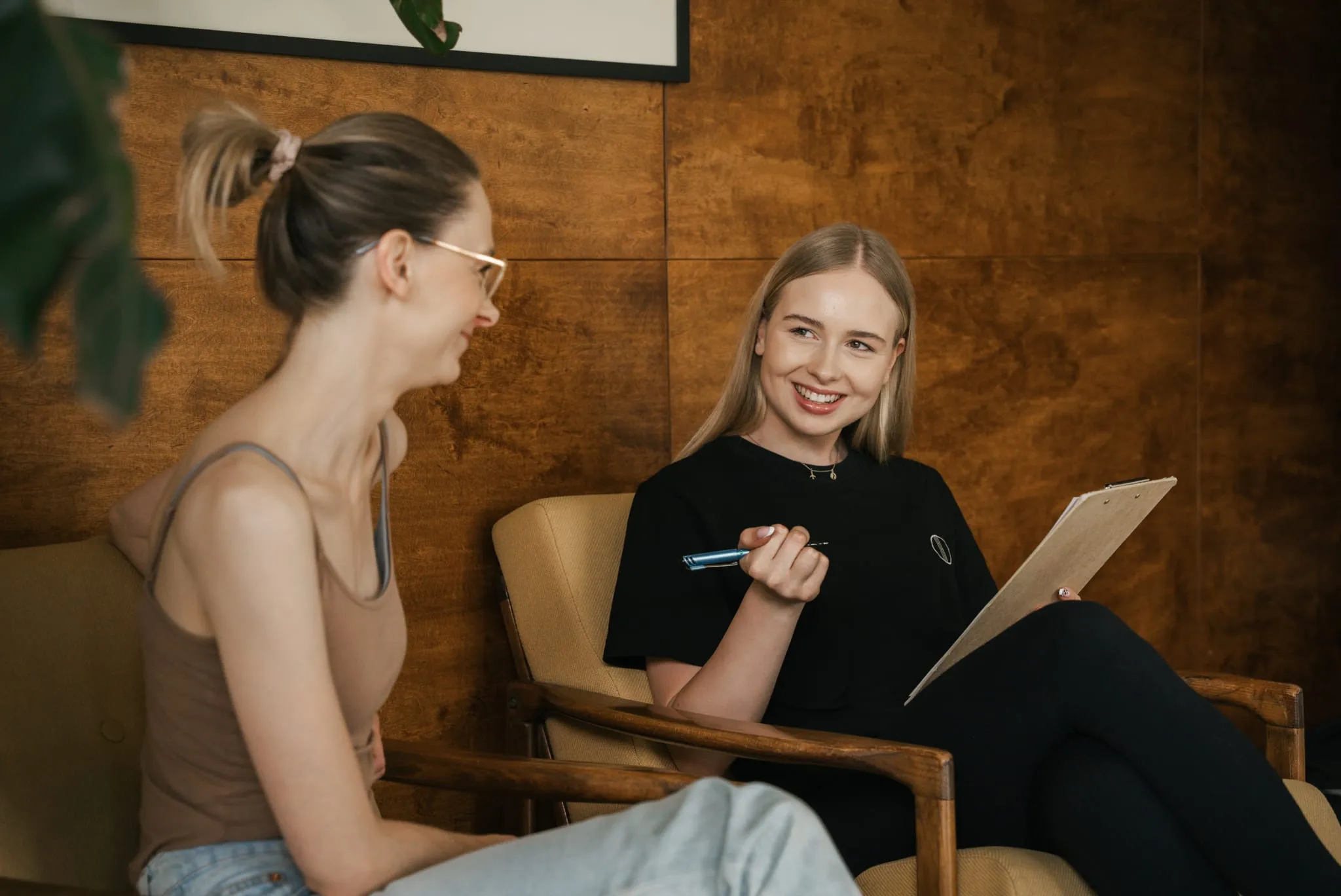 Trenerka z kartą wywiadu rozmawia z klientką w fotelach przy drewnianej ścianie