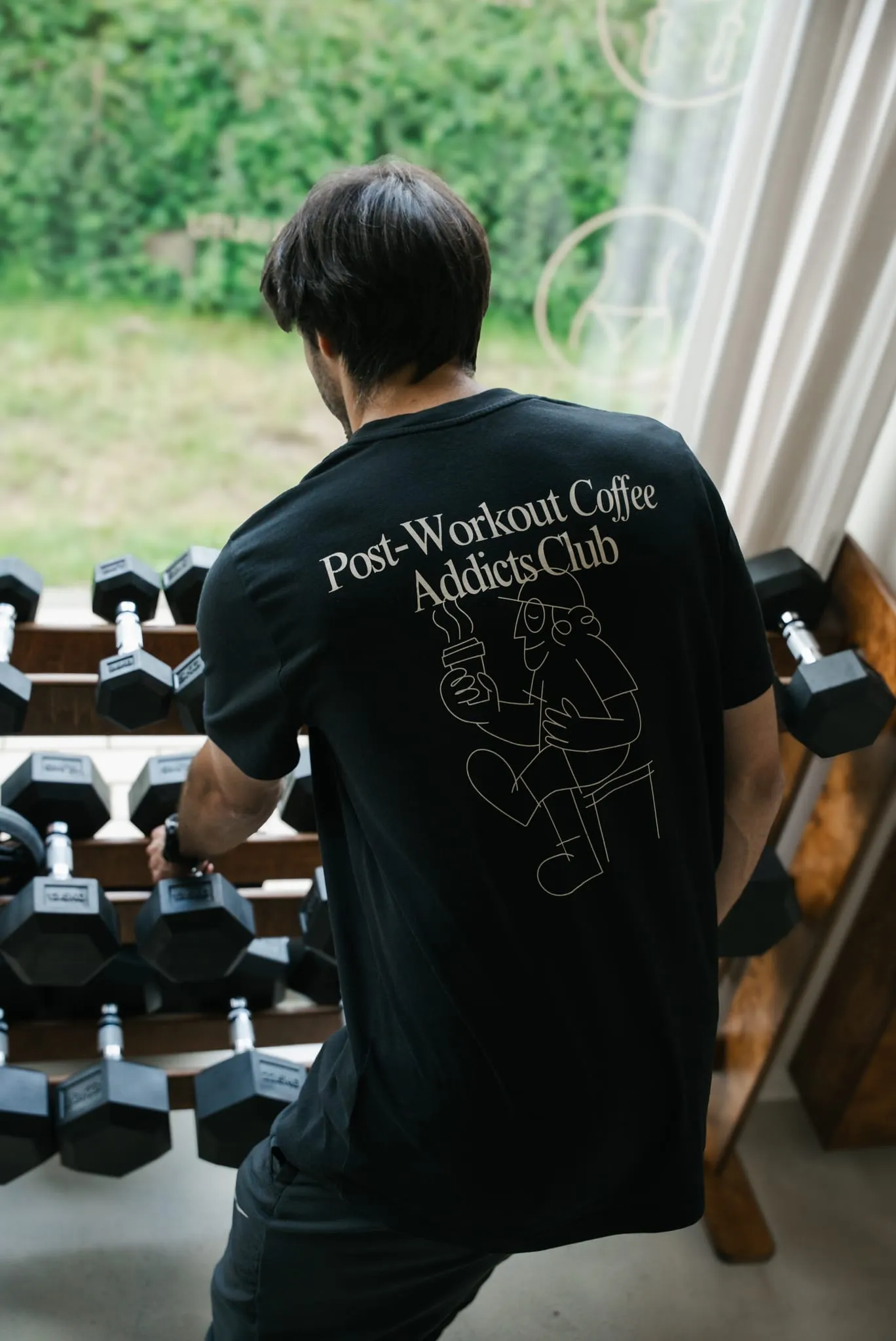 Mężczyzna w czarnym T-shircie WORK IT przy stojaku z hantlami, tyłem do obiektywu