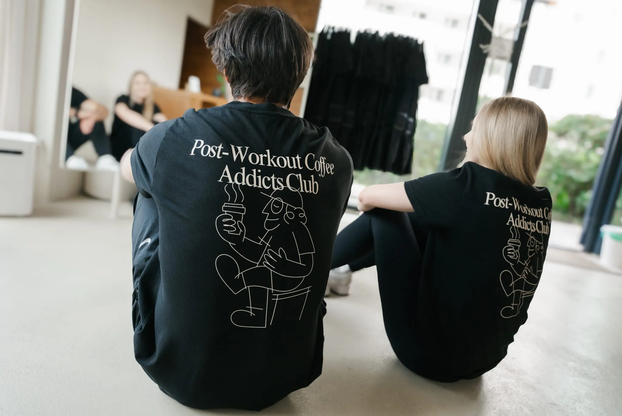 Dwie osoby w T-shirtach WORK IT siedzą na podłodze studia, tyłem do obiektywu
